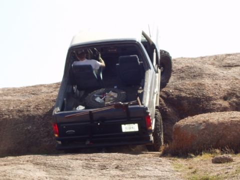 1986 Ford Bronco - A black Ford Bronco navigates a rocky terrain, viewed from the rear with its spare tire and jack loaded.