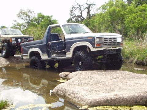 1986 Ford Bronco - Two modified off-road vehicles navigate through shallow water and rocks.