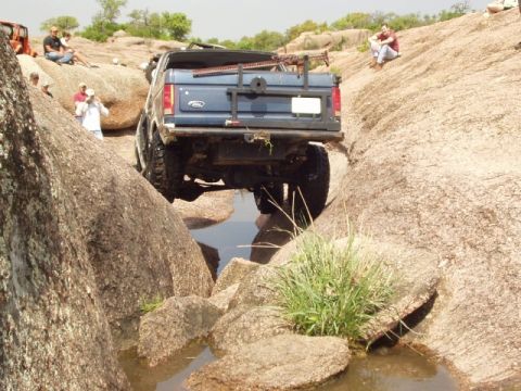 1986 Ford Bronco - A blue Ford Bronco tackles a challenging off-road course with large rocks and water obstacles.