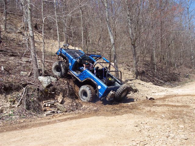 Blue buggy with roll cage going up a steep, wooded hill. One front wheel is off the ground