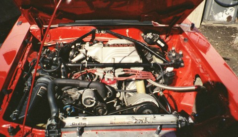 1993 Ford Mustang - The engine bay of a red 1993 Ford Mustang Cobra R is visible with the hood raised, revealing the V8 engine and associated components.