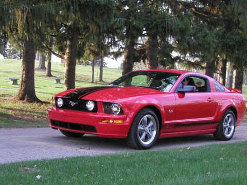 2005 Ford Mustang - A red 2005 Ford Mustang GT coupe is parked on a road with trees in the background.