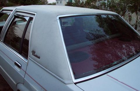 1985 Ford Crown Victoria LTD - The rear passenger side of a sedan is shown, with a vinyl roof and a reflection of trees in the window.