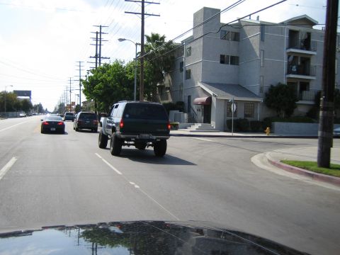 1990 Ford Bronco - black lifted SUV drives away down a tree-lined residential street past apartment buildings, photographed from a following car.