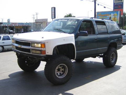1990 Ford Bronco - heavily lifted green-and-white Chevrolet SUV with a bowtie grille emblem and large tires parked in front of storefronts.