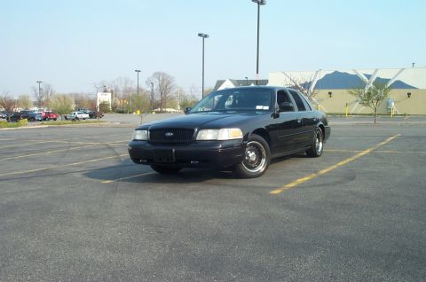 1999 Ford Crown Victoria - Black Ford Crown Victoria sedan sitting alone in an empty asphalt lot, photographed from the driver's-side three-quarter front under a clea…