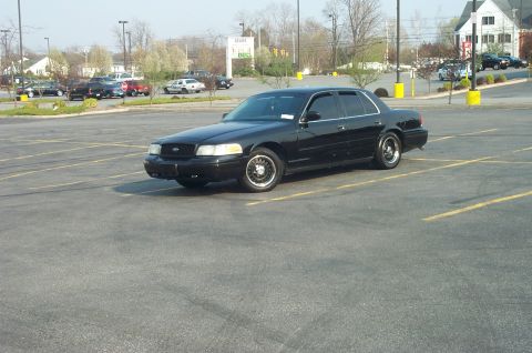 1999 Ford Crown Victoria - Black Ford Crown Victoria sedan parked at an angle across empty striped parking spaces, shot from the driver's-side three-quarter front. It…