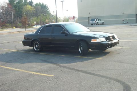 1999 Ford Crown Victoria - A black sedan with aftermarket wheels is parked in a lot, showing its front and side profile.