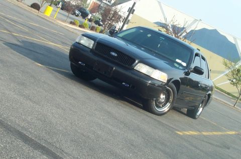 1999 Ford Crown Victoria - Tilted low-angle three-quarter front shot of a black Ford Crown Victoria in an empty parking lot, showing the blacked-out egg-crate grille…