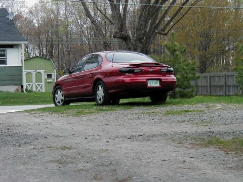 1997 Ford Taurus - Rear view of a red 1997 Ford Taurus sedan parked in a gravel lot with a wooden fence in the background.