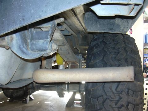 1991 Ford Bronco - Underside view of a lifted truck on a jack stand, showing a large cylindrical steel muffler mounted crosswise, the frame rail, driveshaft a…