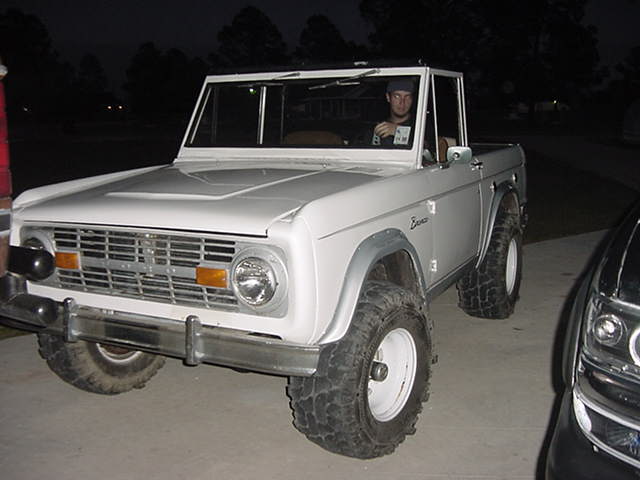 White early Ford Bronco at night, 3/4 front view, big tires, top removed, driver inside.