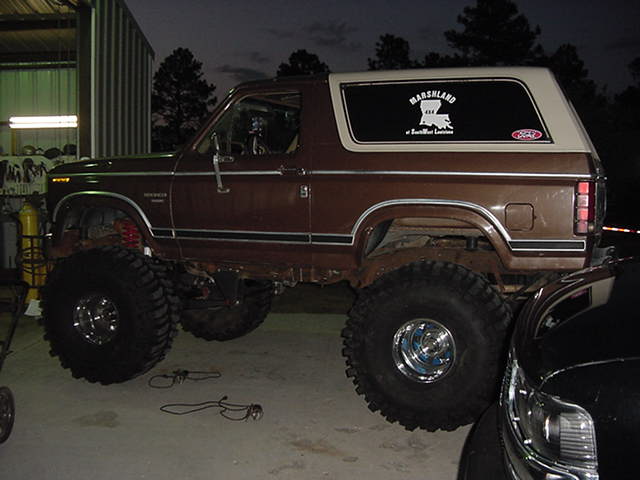 Lifted brown two-door Ford Bronco with huge off-road tires parked at night by a metal shop building