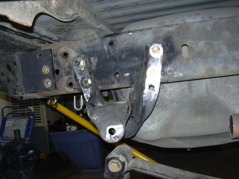 1991 Ford Bronco - Underside view of a truck/SUV frame showing a freshly welded, bare-metal bracket or crossmember mount bolted to the frame rail, with a yell…