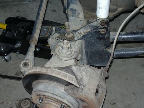 1991 Ford Bronco - Close-up of a front suspension and brake assembly viewed from below, showing a disc brake rotor and caliper, a rusty spindle with threaded…