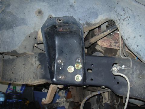 1991 Ford Bronco - Underside view of a vehicle frame crossmember area showing a black-painted steel bracket bolted to the frame rail with several fresh gold-z…