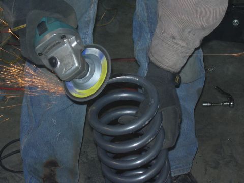 1991 Ford Bronco - A person kneeling in worn blue jeans uses a Makita angle grinder with a flap disc to cut or grind a gray coil spring, throwing bright orang…