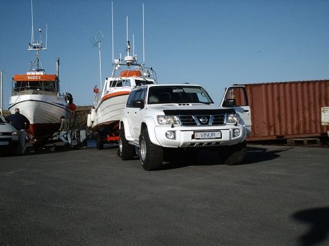 1996 Ford Bronco - A white Nissan Patrol SUV is parked next to a boat trailer in a harbor area, with a shipping container in the background.