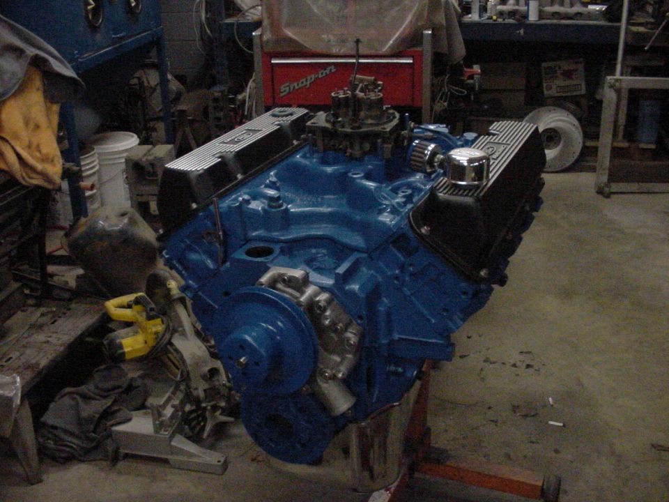 Blue V8 engine with chrome valve covers and carburetor on an engine stand in a workshop