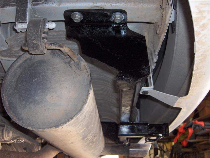 Black bolt-on hitch bracket mounted under white car next to muffler and exhaust hanger