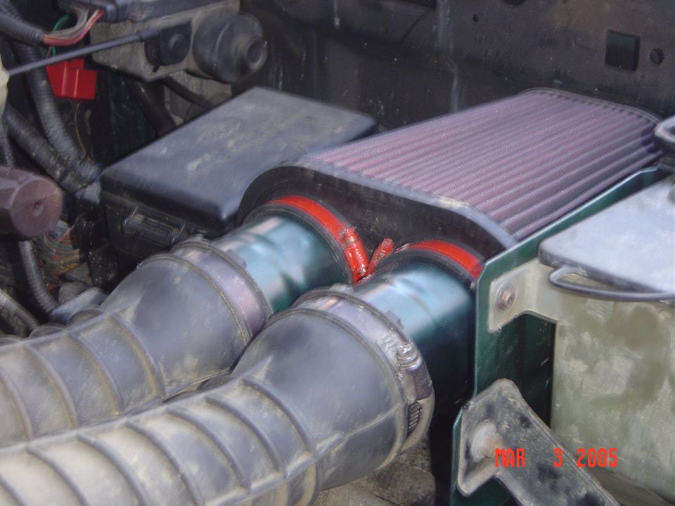 Engine bay with red aftermarket air filter, dual metal intake tubes, red couplers and black corrugated hoses