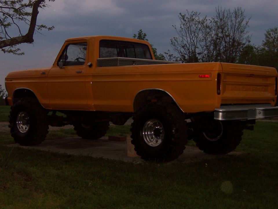 Lifted orange-yellow Ford short-bed pickup on chrome wheels and mud tires at dusk