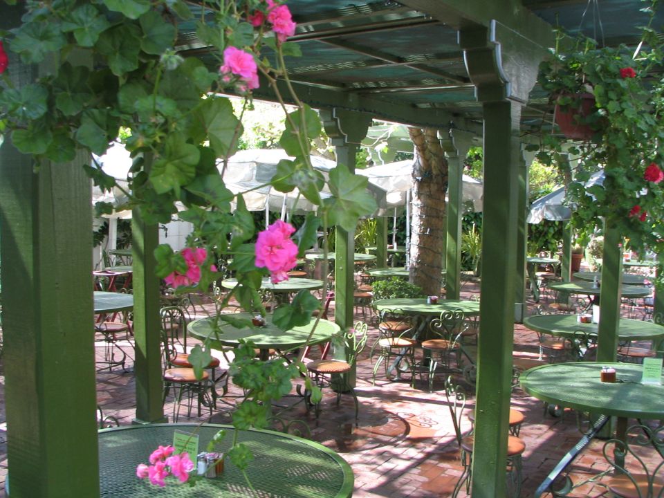 Outdoor cafe seating area with green tables and chairs, pink flowers, and a Supermotors.net watermark