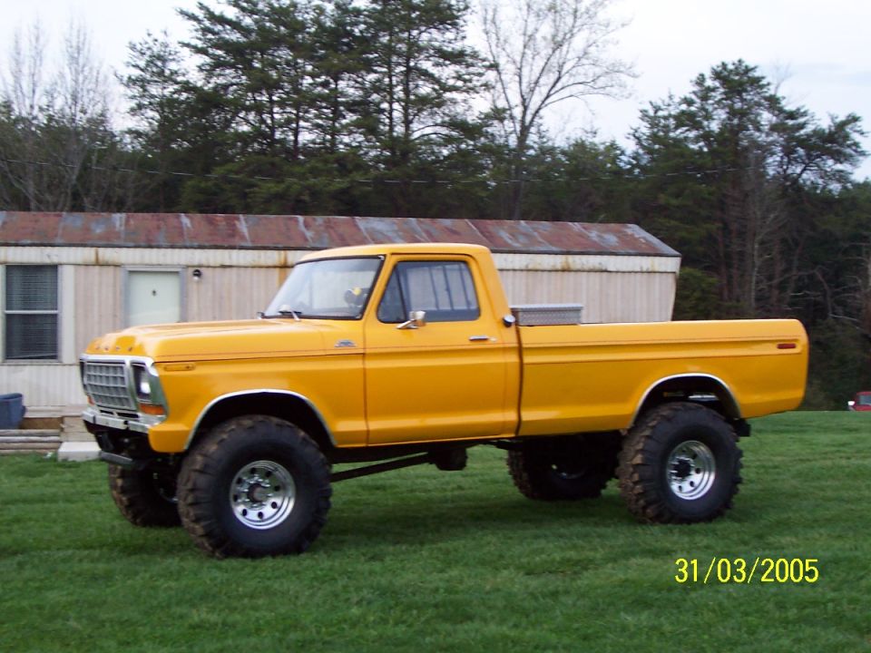 Lifted yellow-orange Ford 4x4 pickup on mud tires parked on grass by a single-wide trailer