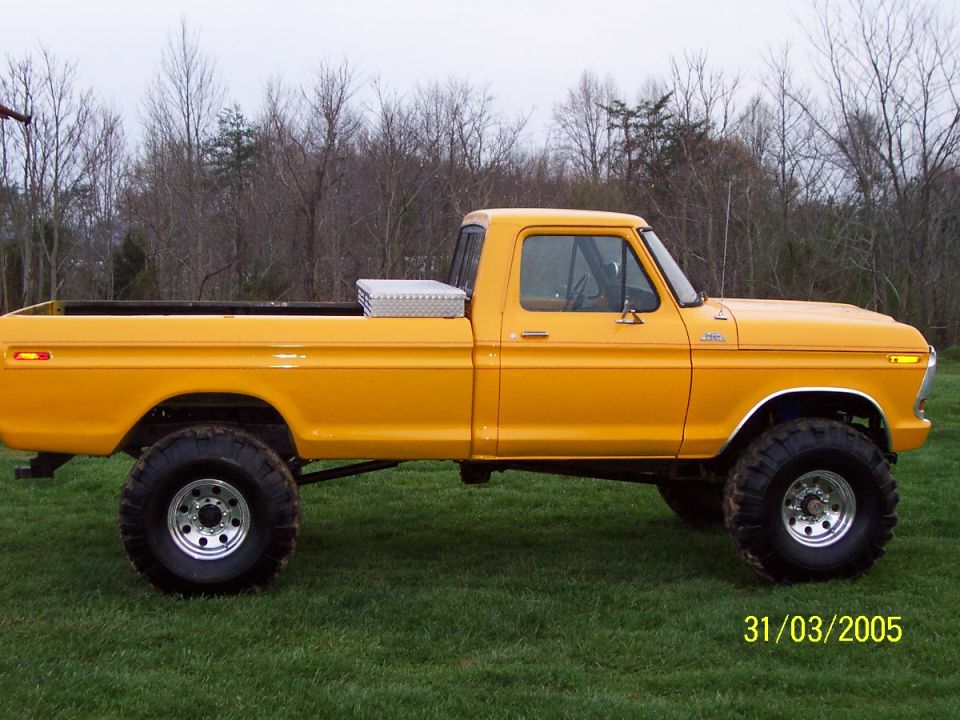 Side profile of a lifted yellow Ford long-bed 4x4 pickup on grass with bare winter trees behind
