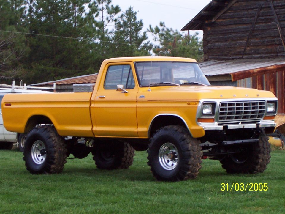 Three-quarter front of a lifted orange-yellow Ford 4x4 pickup parked near old log outbuildings