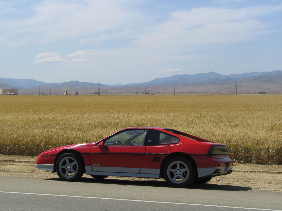 Red 1986 Pontiac Fiero parked on the side of a road with a field of wheat and mountains in the background