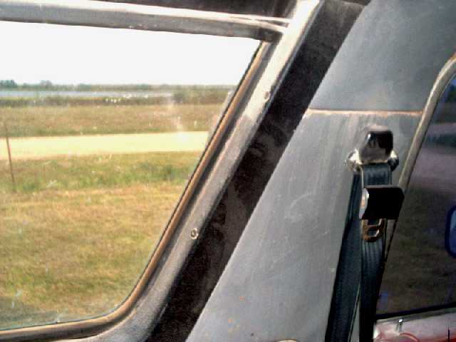Interior view of B-pillar and seat belt hardware next to side window, grassy field visible outside