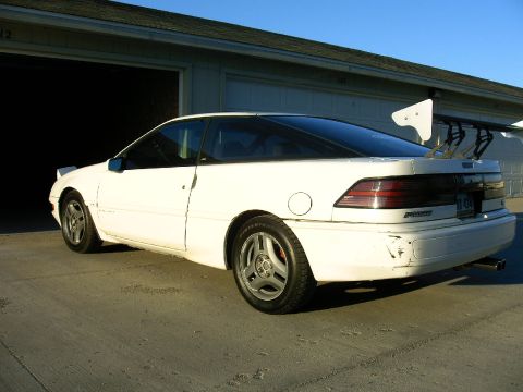 1992 Ford Probe - A white 1992 Ford Probe is parked outside a garage, showing off its large rear spoiler and some damage to the bumper.