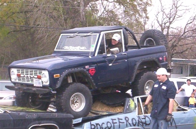 Blue early Ford Bronco climbing a dirt obstacle at a show, driver in helmet, spectators around