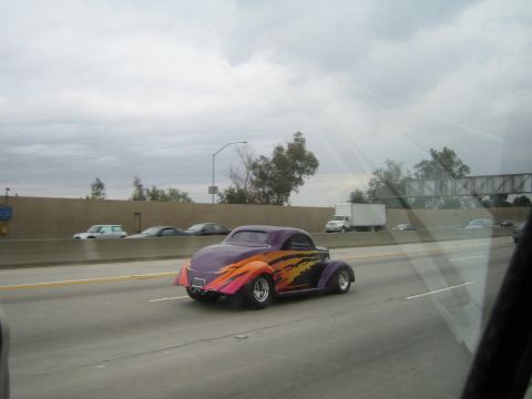 1990 Ford Bronco - purple street-rod coupe with orange flame graphics cruises a freeway, photographed from another vehicle.
