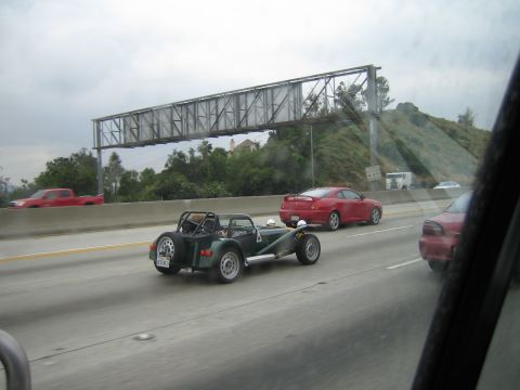 1990 Ford Bronco - green open-wheel Lotus Seven-style roadster travels a freeway under an overhead sign gantry beside a red coupe.