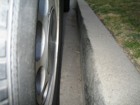 1985 Ford Mustang - Extreme low-angle close-up along a curb of a silver five-spoke alloy wheel with a Ford center cap, showing the tire sidewall and the car's…
