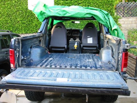 1985 Ford Bronco - The interior and bed of a 1985 Ford Bronco are visible, with a green tarp covering the roof area.
