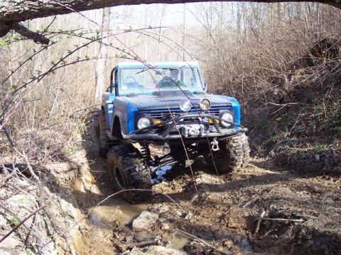 1974 Ford Bronco - A modified 1974 Ford Bronco tackles a challenging muddy trail in a wooded area.
