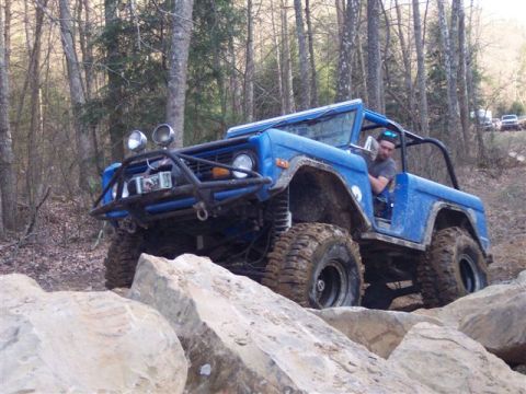 1974 Ford Bronco - A blue SUV is shown off-roading through a rocky, wooded area. The vehicle has large tires and a modified bumper.