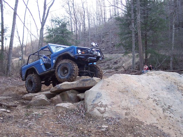 Blue 1974 Ford Bronco with roll cage and off-road tires climbing over large rocks