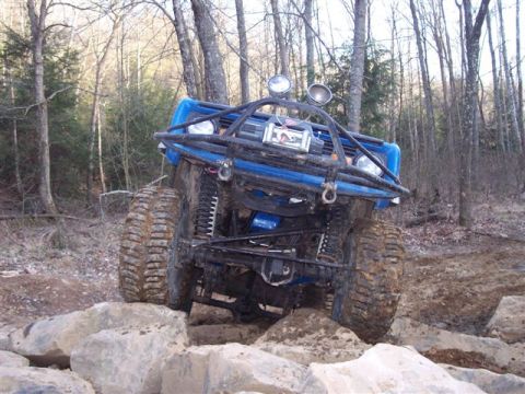1974 Ford Bronco - A modified blue SUV navigates a rocky trail in a forest setting, its off-road capabilities.