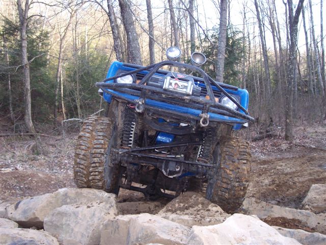 Blue off-road SUV with large tires crawling over rocks in a wooded area. It has a winch and extra lights on the front