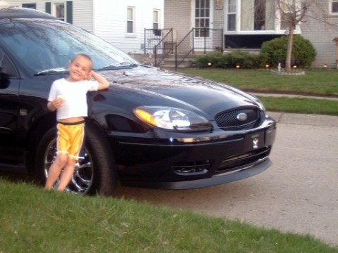 2004 Ford Taurus - A young boy poses with a black sedan parked on a residential street.