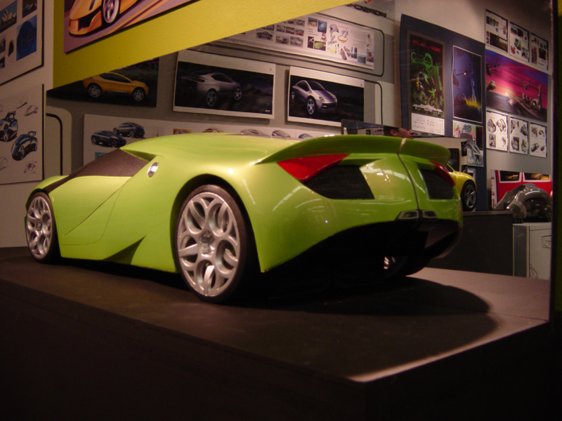 Green concept supercar model rear three-quarter on stand, walls covered with car renderings