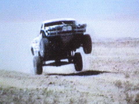 1990 Ford Bronco - grainy screen-captured photo of an off-road race truck catching air over desert terrain.