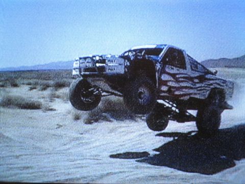 1990 Ford Bronco - blurry screen photo of a trophy-style off-road truck mid-jump over a sandy desert berm.