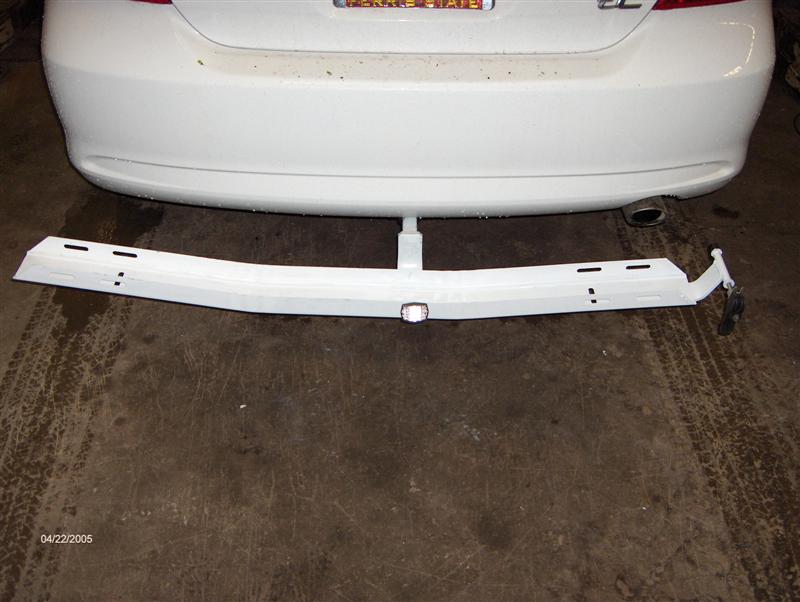 White trailer hitch crossmember on shop floor behind a white coupe's rear bumper