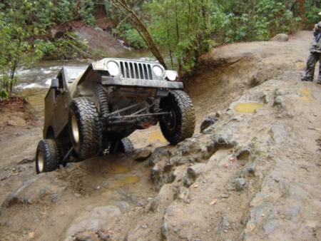 1989 Ford Bronco - A modified Jeep Wrangler TJ tackles a challenging off-road course, demonstrating its capabilities on a wet, uneven terrain.