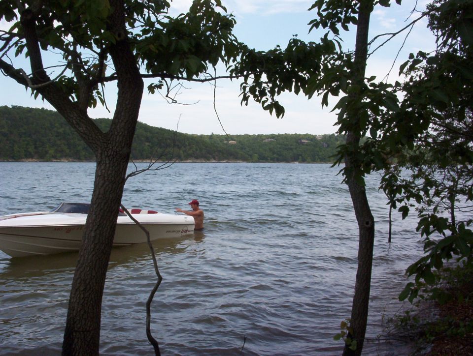 White speedboat with red interior beached along a wooded lakeshore, person standing in water nearby
